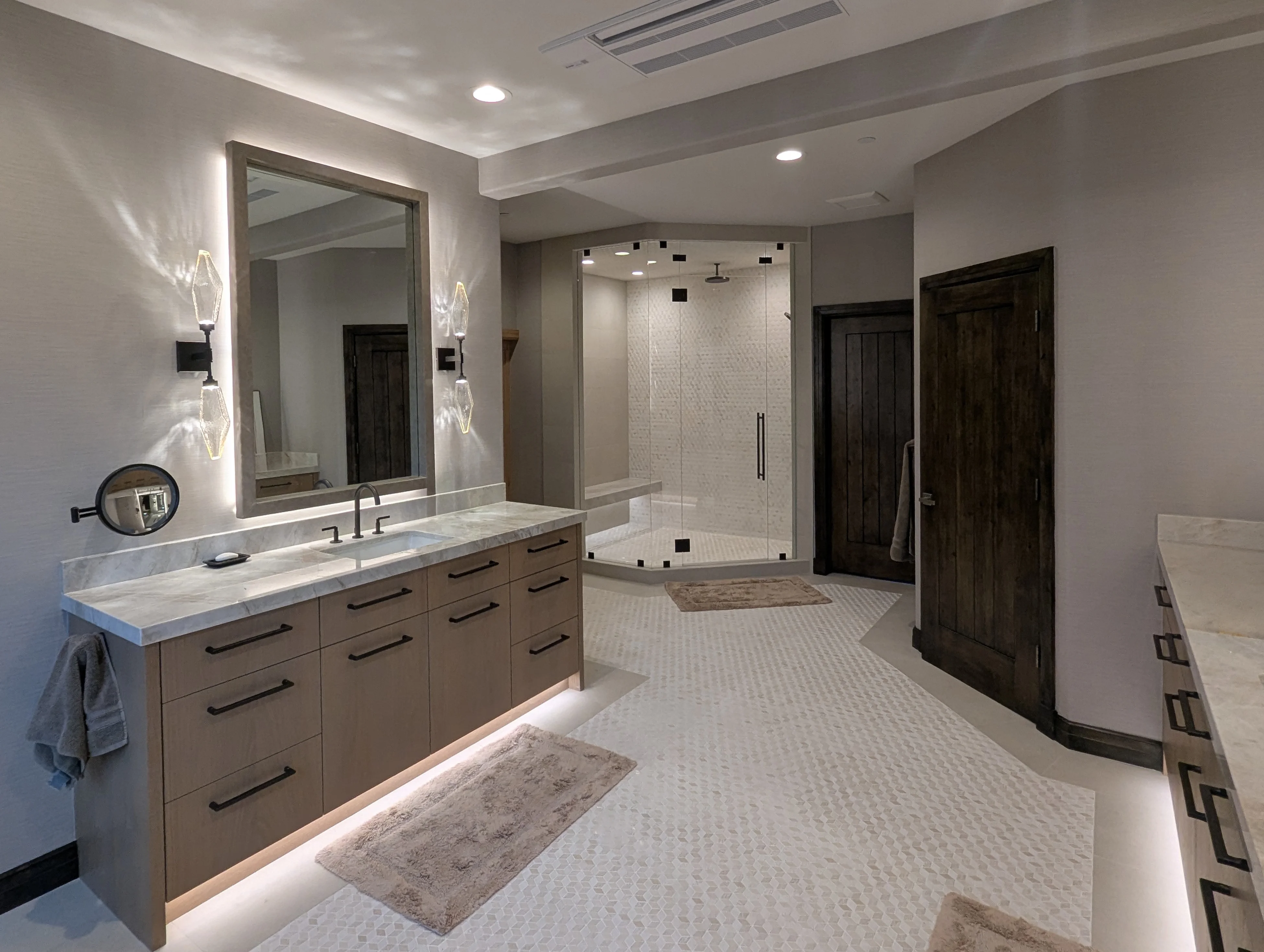 Wide view of a primary bathroom showing both vanities and the glass corner shower