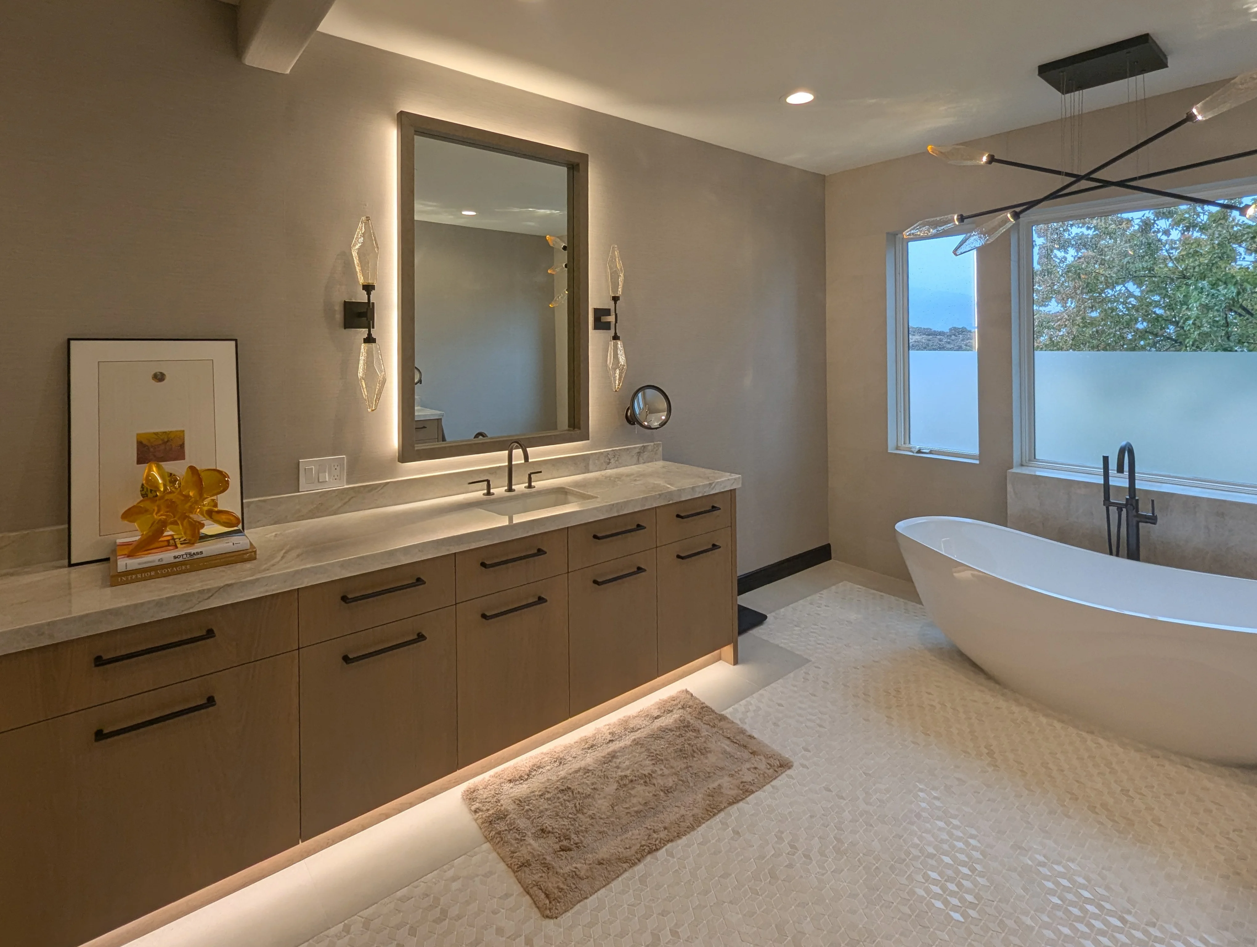 Floating white oak vanity with a backlit mirror, and a freestanding tub by the window